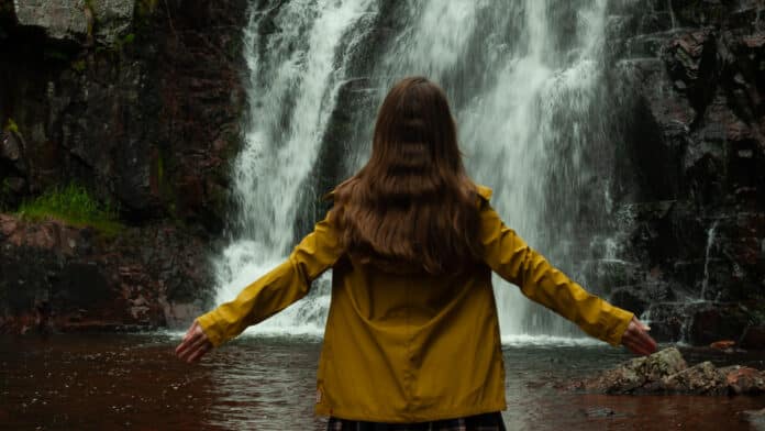 Lisah Silfwer in a dirty yellow raincoat in front of a waterfall.
